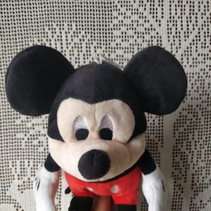 Puede incluir: Un juguete de peluche de Mickey Mouse con orejas negras, nariz negra y pantalones cortos rojos con lunares blancos. El juguete tiene una cara blanca con ojos negros y una gran sonrisa.