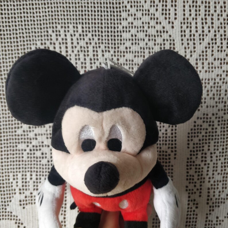Puede incluir: Un juguete de peluche de Mickey Mouse con orejas negras, nariz negra y pantalones cortos rojos con lunares blancos. El juguete tiene una cara blanca con ojos negros y una gran sonrisa.