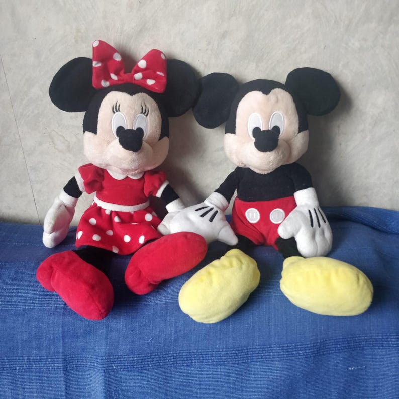 Puede incluir: Un par de peluches de Mickey Mouse y Minnie Mouse. Mickey lleva pantalones cortos negros con lunares rojos y zapatos amarillos. Minnie lleva un vestido rojo con lunares blancos y zapatos rojos. Ambos tienen orejas negras y guantes blancos.