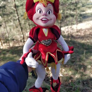 May include: A red plush doll with a yellow bow and a heart-shaped design on its chest. The doll is wearing red and gold shoes and has a white face with painted features.