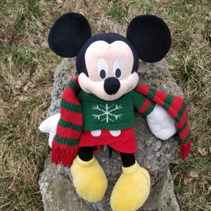 Peut inclure: Un Mickey Mouse en peluche portant un pull vert avec un flocon de neige et une &eacute;charpe ray&eacute;e rouge et verte.