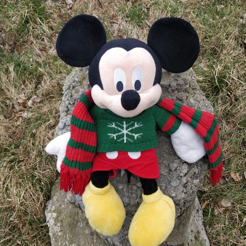 Peut inclure: Un Mickey Mouse en peluche portant un pull vert avec un flocon de neige et une &eacute;charpe ray&eacute;e rouge et verte.