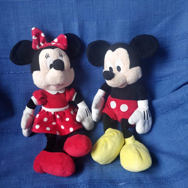 Puede incluir: Un Mickey Mouse de peluche rojo y negro y una Minnie Mouse de peluche roja y blanca. Ambos est&aacute;n de pie sobre una superficie azul.