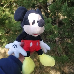 Puede incluir: Un juguete de peluche de Mickey Mouse con orejas negras, un cuerpo rojo y negro y zapatos amarillos. El juguete est&aacute; sentado en una mano.