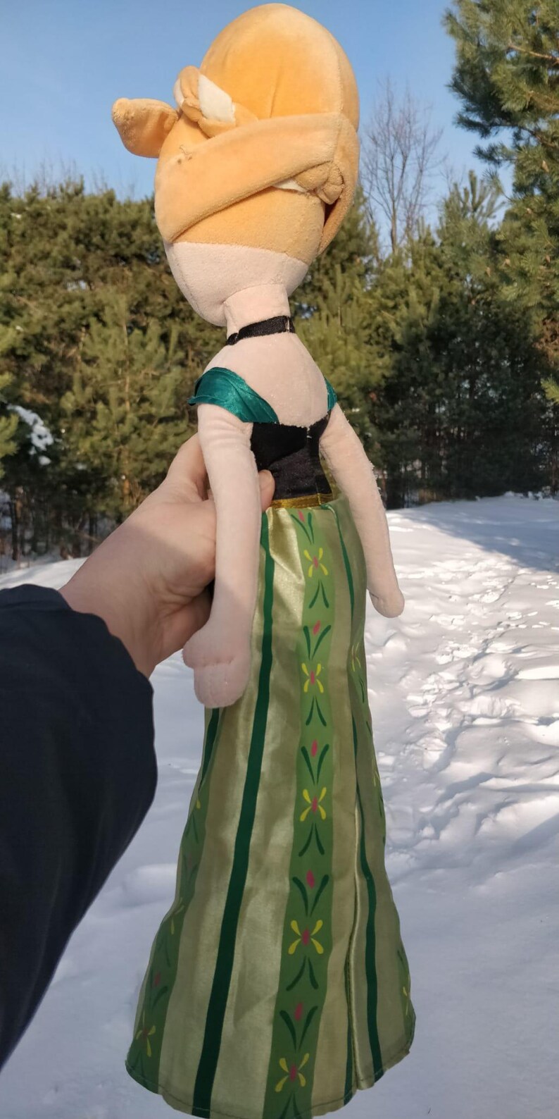 Op de afbeelding: Een pluche pop van een personage uit een populaire animatiefilm. De pop heeft een lichtbruin hoofd met een blond haarstuk, een zwarte choker en een groene en zwarte jurk met een groene en gouden rok. De pop wordt voor een besneeuwde achtergrond gehouden.