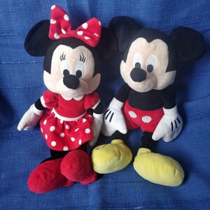 Puede incluir: Un conjunto de juguetes de peluche de Mickey Mouse y Minnie Mouse. Mickey lleva pantalones cortos rojos y zapatos amarillos. Minnie lleva un vestido rojo con lunares blancos y zapatos rojos.