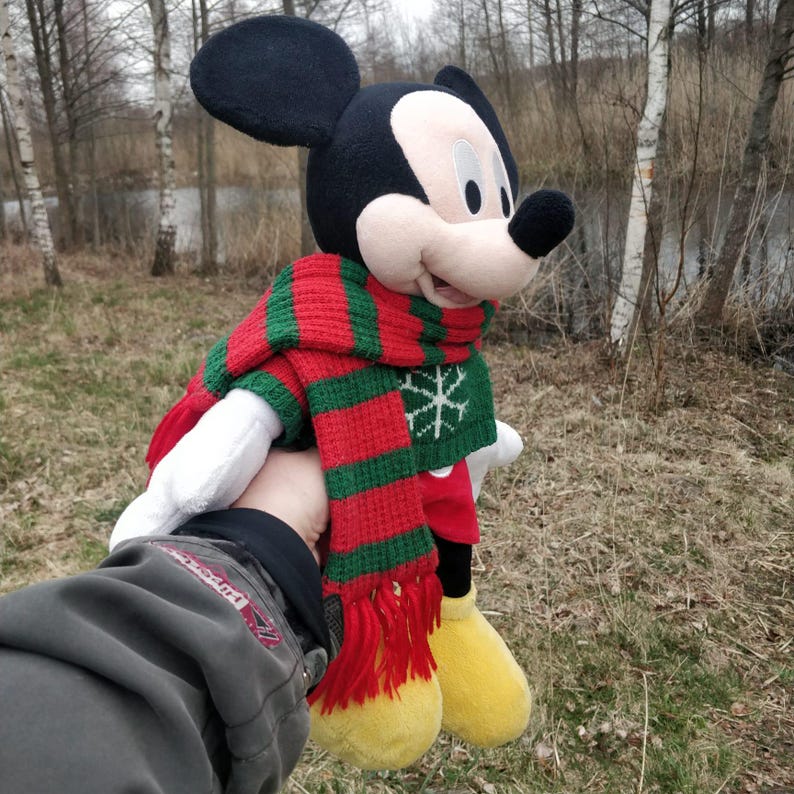 Peut inclure: Un jouet en peluche Mickey Mouse portant une &eacute;charpe ray&eacute;e rouge et verte avec un motif flocon de neige. Le jouet est tenu par une main portant une veste verte.