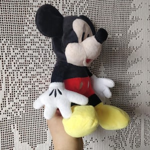 Puede incluir: Un juguete de peluche de Mickey Mouse con pelaje negro, pantalones cortos rojos y zapatos amarillos. El juguete tiene una gran sonrisa y levanta las manos.