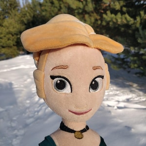 Op de afbeelding: Pluche pop van een blonde vrouw met een gele hoed en een zwarte choker ketting met een gouden hanger. De pop heeft een groene en zwarte jurk met gouden rand. De pop staat tegen een besneeuwde achtergrond met groene bomen.