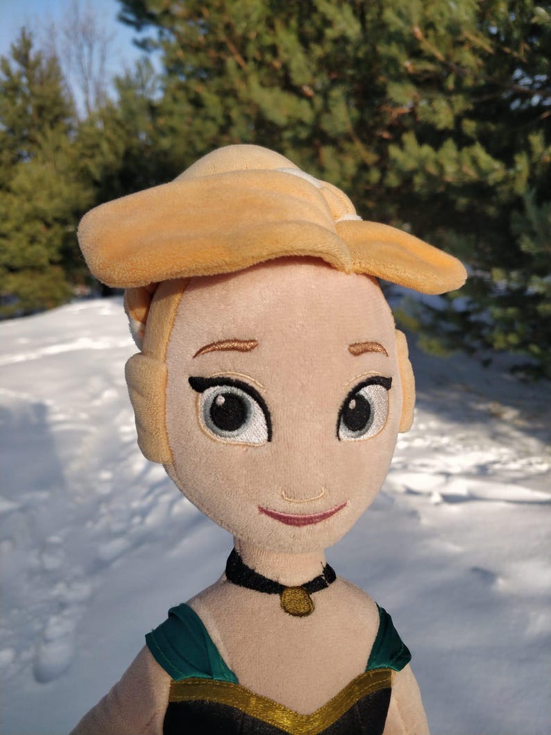 Op de afbeelding: Pluche pop van een blonde vrouw met een gele hoed en een zwarte choker ketting met een gouden hanger. De pop heeft een groene en zwarte jurk met gouden rand. De pop staat tegen een besneeuwde achtergrond met groene bomen.