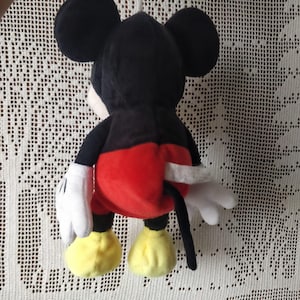 Puede incluir: Un juguete de peluche Mickey Mouse negro y rojo con una cola blanca y zapatos amarillos. El juguete est&aacute; sentado sobre una superficie de crochet blanco.