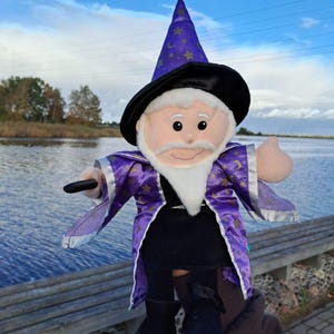 May include: A plush wizard doll with a white beard, black boots, and a purple and gold robe. The wizard wears a black hat with a purple top adorned with gold stars and moons. The doll is holding a black wand.