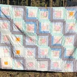 Könnte beinhalten: Eine Patchwork-Decke mit einem karierten Muster in Schwarz und Weiß. Die Decke besteht aus verschiedenen Stoffen in Blautönen, Rosa und Weiß. Die Decke hängt an einer Wäscheleine.