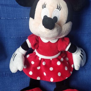 Puede incluir: Un vestido de peluche rojo con lunares blancos y un lazo rojo en la cabeza. El juguete de peluche es un personaje de dibujos animados con orejas negras y una nariz negra. El juguete est&aacute; de pie sobre una superficie azul.