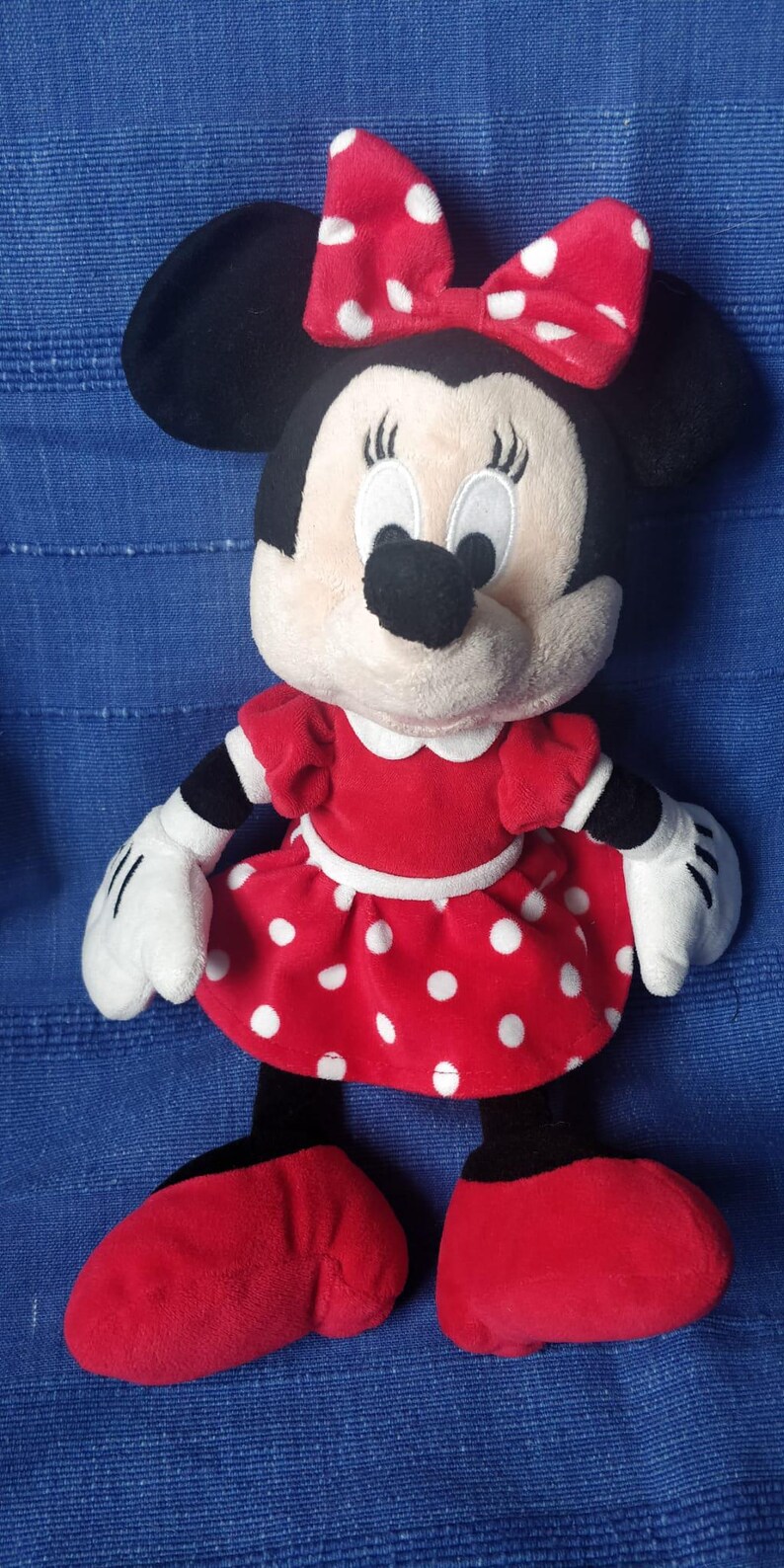 Puede incluir: Un vestido de peluche rojo con lunares blancos y un lazo rojo en la cabeza. El juguete de peluche es un personaje de dibujos animados con orejas negras y una nariz negra. El juguete est&aacute; de pie sobre una superficie azul.