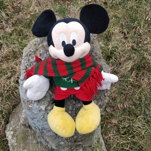 Peut inclure: Un jouet en peluche Mickey Mouse portant une &eacute;charpe ray&eacute;e rouge et verte. Le jouet est assis sur une surface de pierre grise.