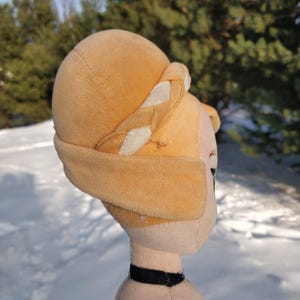 Op de afbeelding: Een pluche pop met een lichtbruin hoofd en een bijpassende hoed met witte accenten. De pop heeft een zwarte choker en een groen en zwart kledingstuk. De achtergrond is een besneeuwd landschap met groenblijvende bomen.