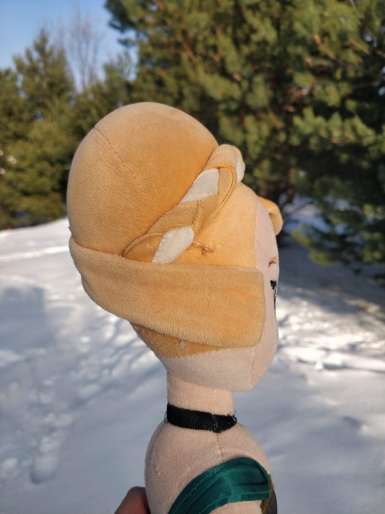 Op de afbeelding: Een pluche pop met een lichtbruin hoofd en een bijpassende hoed met witte accenten. De pop heeft een zwarte choker en een groen en zwart kledingstuk. De achtergrond is een besneeuwd landschap met groenblijvende bomen.