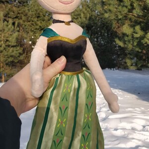 Op de afbeelding: Een pluche pop van een personage met een blonde knot, gekleed in een zwarte en groene jurk. De pop heeft een lachend gezicht en wordt tegen een besneeuwde achtergrond gehouden.