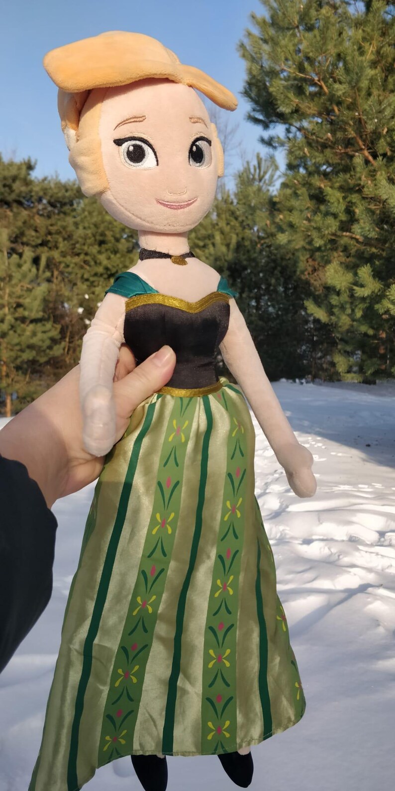 Op de afbeelding: Een pluche pop van een personage met een blonde knot, gekleed in een zwarte en groene jurk. De pop heeft een lachend gezicht en wordt tegen een besneeuwde achtergrond gehouden.