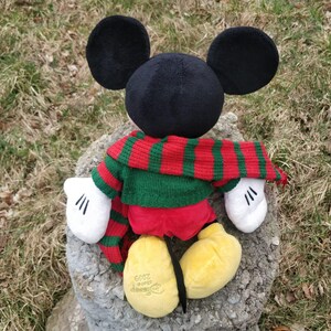 Peut inclure: Un jouet en peluche Mickey Mouse portant une &eacute;charpe ray&eacute;e rouge et verte et un pull vert. Le jouet est assis sur un rocher avec un logo Disney 2002 sur son pied.