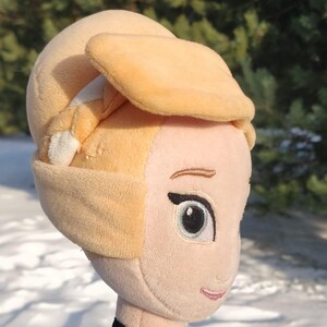Op de afbeelding: Een pluche pop van een prinses met een lichte huidskleur, met een gele hoofdbedekking en een zwarte choker-ketting met een gouden hanger. De pop heeft een groene en zwarte jurk. De achtergrond is een besneeuwd landschap.
