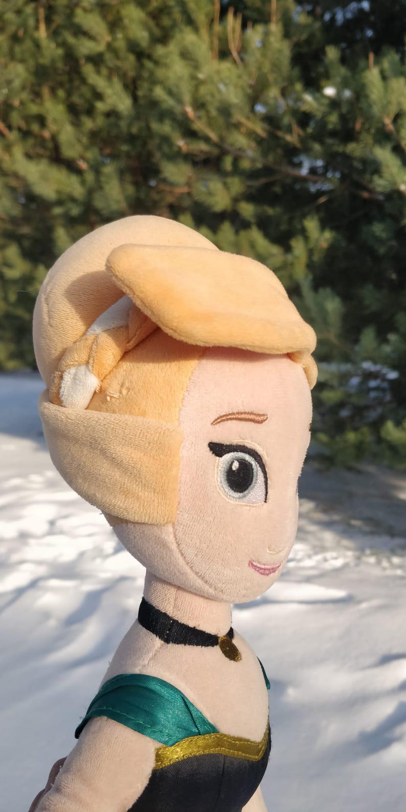 Op de afbeelding: Een pluche pop van een prinses met een lichte huidskleur, met een gele hoofdbedekking en een zwarte choker-ketting met een gouden hanger. De pop heeft een groene en zwarte jurk. De achtergrond is een besneeuwd landschap.