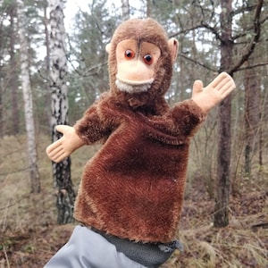 May include: A brown plush monkey puppet with a light tan face, ears, and hands. The puppet has red eyes and is set against a blurred forest background. The monkey's arms are outstretched, giving the impression of a welcoming gesture.