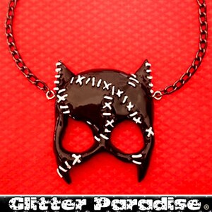 May include: Black catwoman mask necklace with a silver chain. The mask has white stitching details and is on a red background. The text "Glitter Paradise" is at the bottom of the image.
