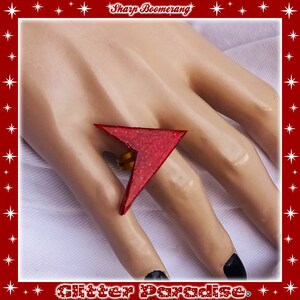May include: A red glitter ring shaped like a sharp boomerang. The ring is worn on a finger of a hand.