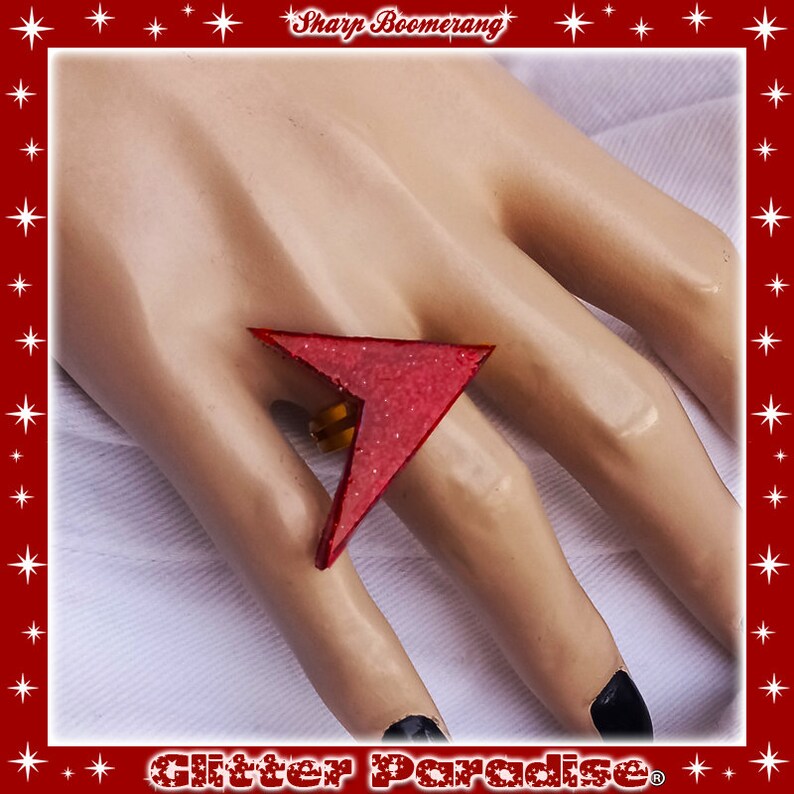 May include: A red glitter ring shaped like a sharp boomerang. The ring is worn on a finger of a hand.