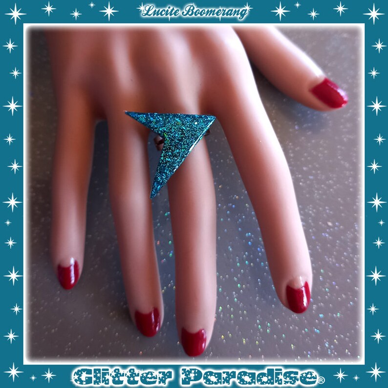 May include: A turquoise glitter ring shaped like a triangle with a pointed tip. The ring is on a hand with red nail polish.
