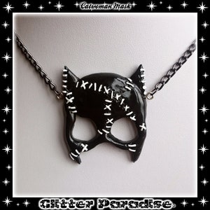 May include: Black and white catwoman mask necklace with a chain. The mask has white stitching details.