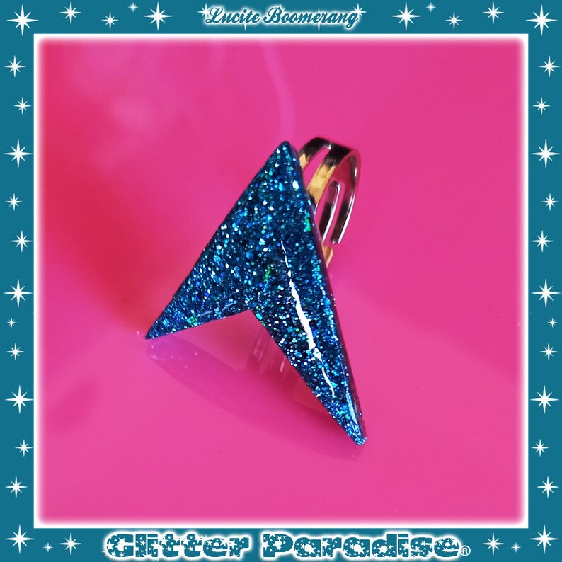 May include: A blue glitter arrow ring with a silver band. The ring is on a pink background.
