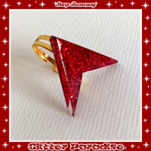 May include: A red glitter arrow ring with a gold band. The ring is a statement piece and would be a great addition to any jewelry collection.