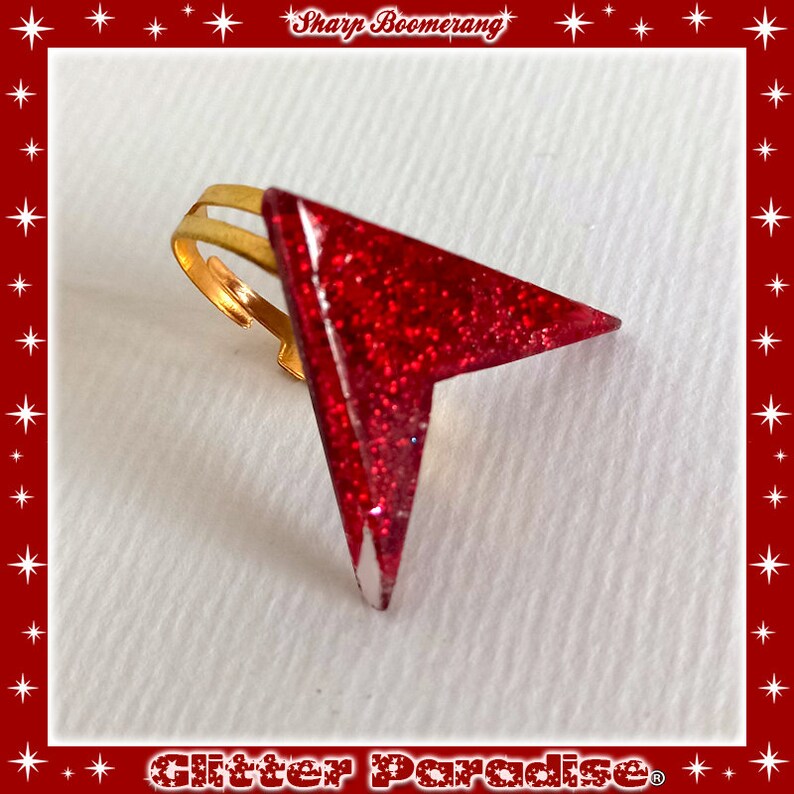May include: A red glitter arrow ring with a gold band. The ring is a statement piece and would be a great addition to any jewelry collection.