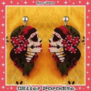 Peut inclure: Une paire de boucles d'oreilles pendantes avec un motif de crâne de sucre du Jour des Morts avec des accents floraux. Les boucles d'oreilles sont en bois et ont des crochets en métal argenté.
