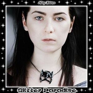 May include: A woman with long black hair wearing a black necklace with a black cat mask pendant. The necklace is made of metal and has a shiny finish. The woman is looking at the camera with a serious expression.