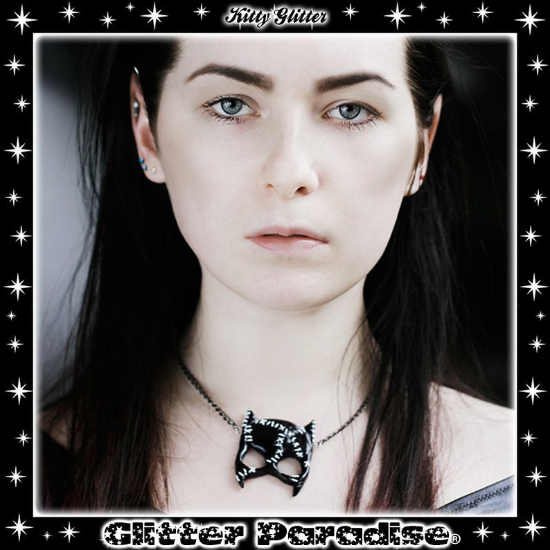 May include: A woman with long black hair wearing a black necklace with a black cat mask pendant. The necklace is made of metal and has a shiny finish. The woman is looking at the camera with a serious expression.
