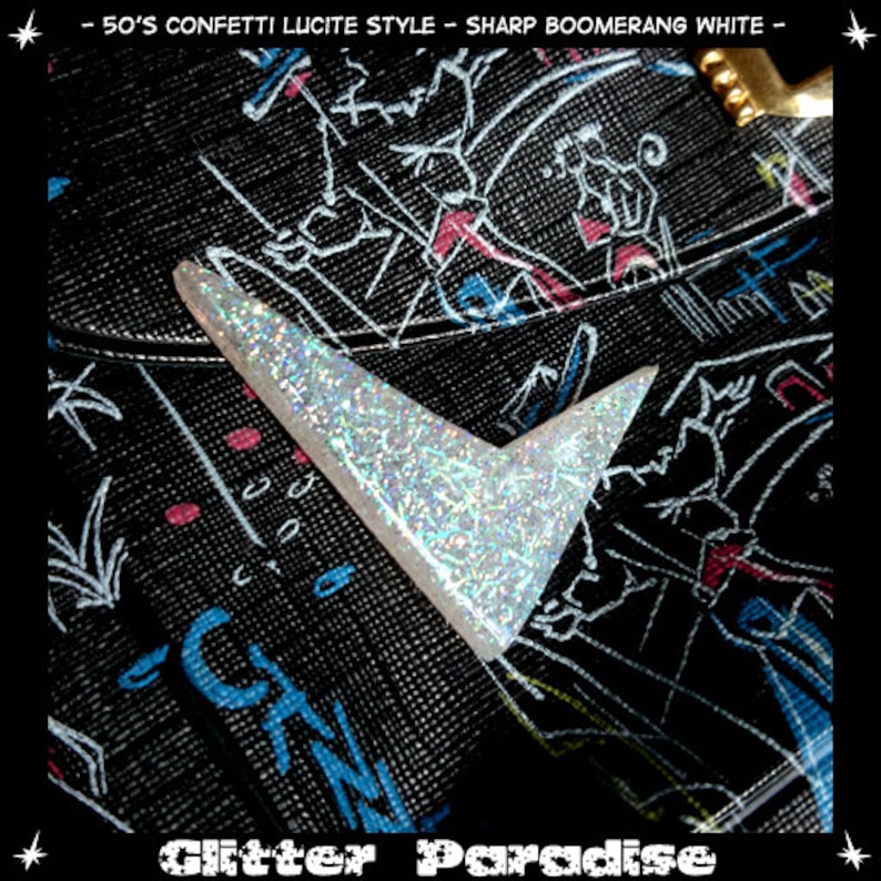 May include: A white glitter boomerang-shaped lucite charm with a black background featuring white, pink, and blue drawings. The text "Glitter Paradise" is visible at the bottom of the image.