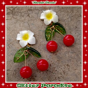 May include: A pair of earrings featuring white plumeria flowers with yellow centers, green leaves, and red cherry accents. The earrings are labeled "Hawaiian Cherries" and "Glitter Paradise".