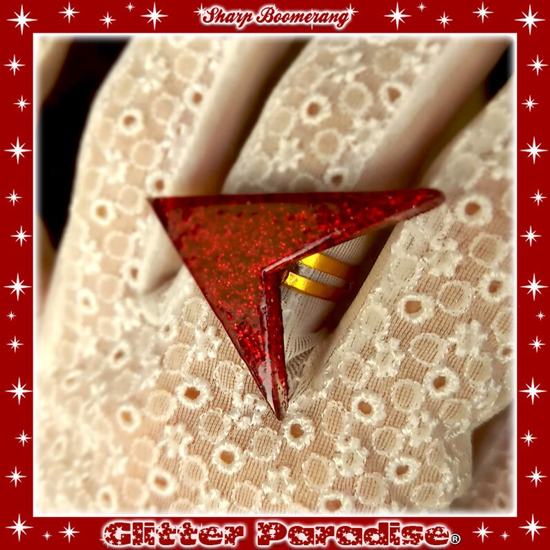 May include: A red glitter boomerang ring with gold accents. The ring is shaped like a triangle and has a sharp point. The ring is on a white lace background.