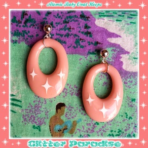 Peut inclure: Une paire de boucles d'oreilles créoles ovales roses avec des accents d'étoiles blanches. Les boucles d'oreilles sont sur un fond de tissu à motifs verts et violets.