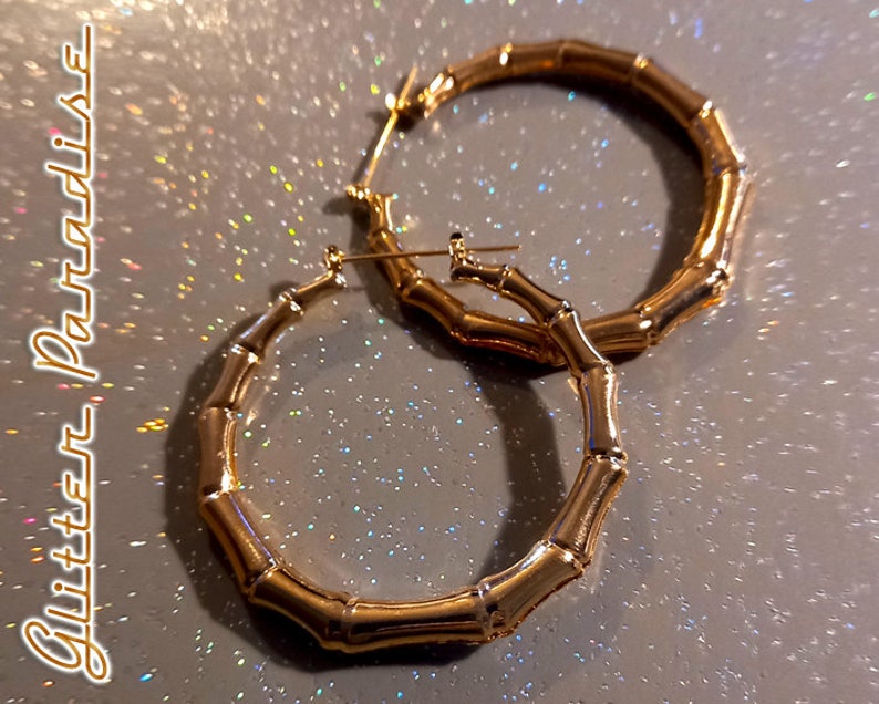 K&ouml;nnte beinhalten: Ein Paar goldfarbene Creolen mit einer strukturierten Bambus-Optik. Die Ohrringe haben eine kreisf&ouml;rmige Form mit einem segmentierten Aussehen, das an Bambusstangen erinnert. Der Text "Glitter Paradise" ist auf der linken Seite zu sehen.