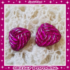 May include: A pair of pink glitter earrings shaped like a knot. The earrings are on a white fuzzy surface.