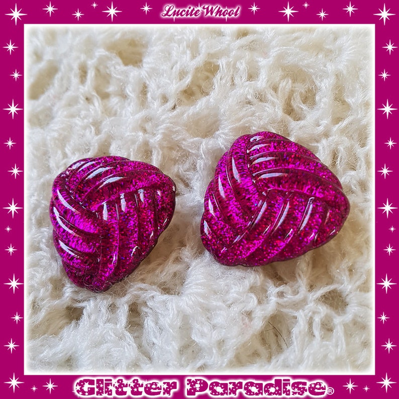 May include: A pair of pink glitter earrings shaped like a knot. The earrings are on a white fuzzy surface.