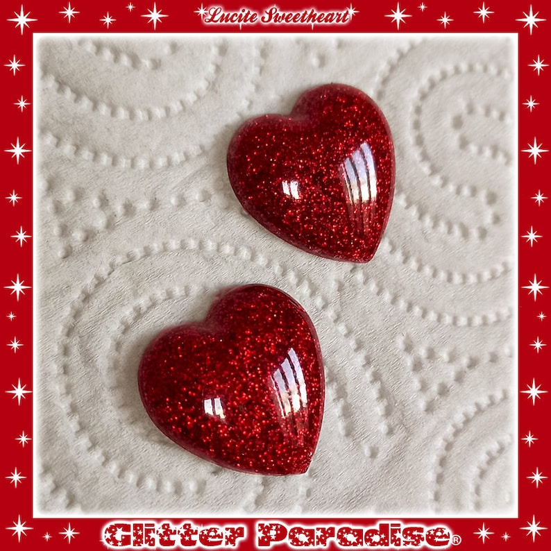 May include: Two red glitter heart-shaped resin cabochons. The hearts are on a white paper background.