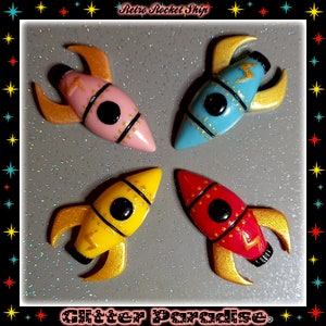 May include: Four miniature rocket ship figurines in pink, blue, yellow, and red with gold accents. The rocket ships have a black circle for a window and a lightning bolt design.
