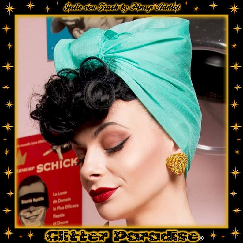 May include: A woman with dark curly hair is wearing a turquoise turban. She has red lipstick and gold earrings. The background is a pink wall with a book on a shelf.