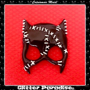 May include: A black and white Catwoman mask with stitched details. The mask is made of a glossy material and has a cat-like shape with pointed ears.